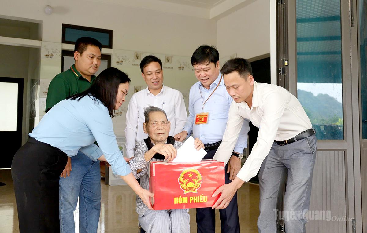 Đồng chí Mai Đức Thông, Ủy viên Ban Chấp hành Đảng bộ tỉnh, Giám đốc Báo và phát thanh, truyền hình Tuyên Quang, Ủy viên Ban Chỉ đạo Bầu cử tỉnh, Ủy viên Ủy ban Bầu cử tỉnh và tổ bầu cử thôn Phúc Ninh mang hòm phiếu phụ đến gia đình cử tri Lý Văn Hoa, 103 tuổi, dân tộc Dao thanh y để cử tri thực hiện quyền bầu cử. Ảnh: Như Quỳnh.