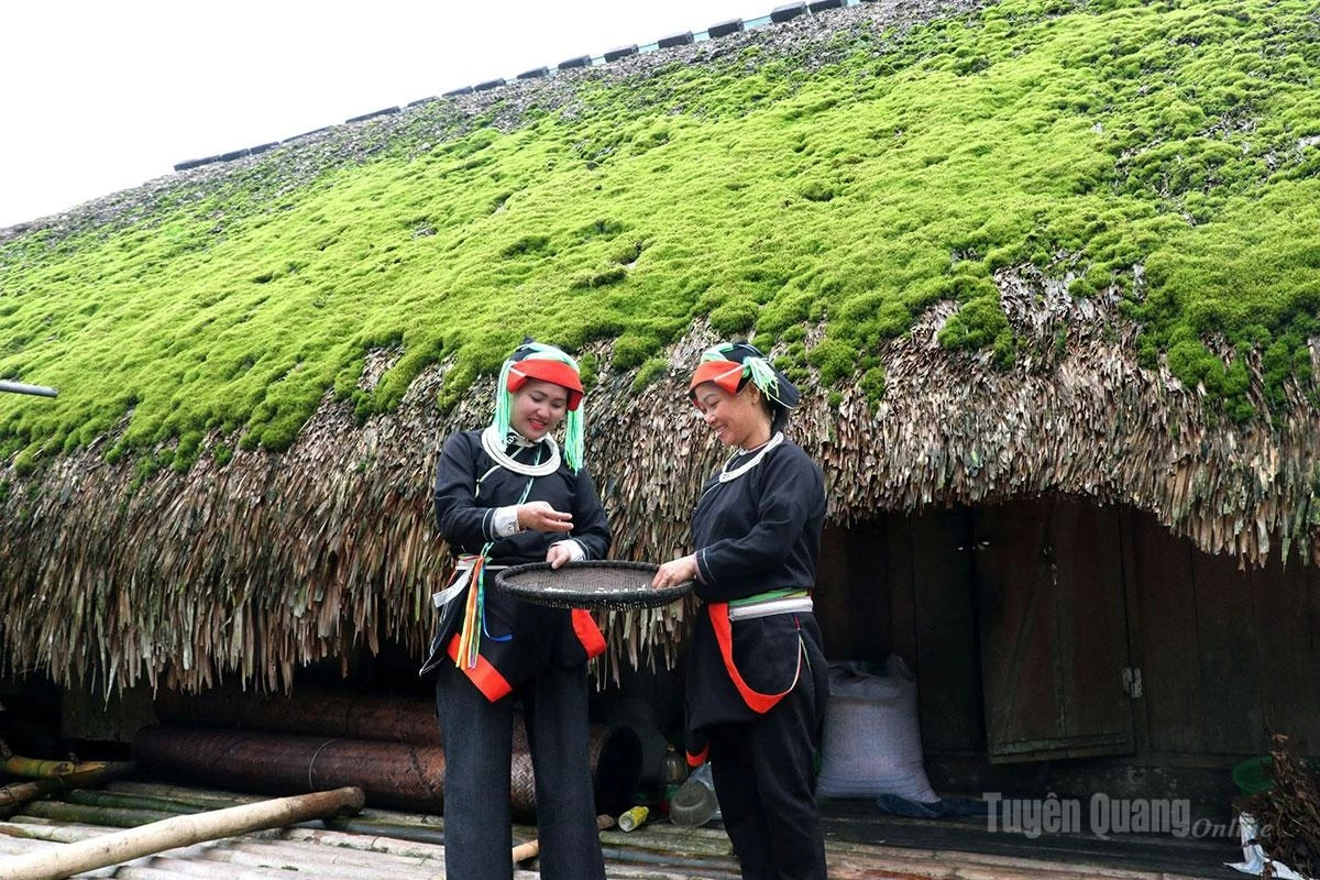 A Dao woman stands beside a traditional moss-covered house.